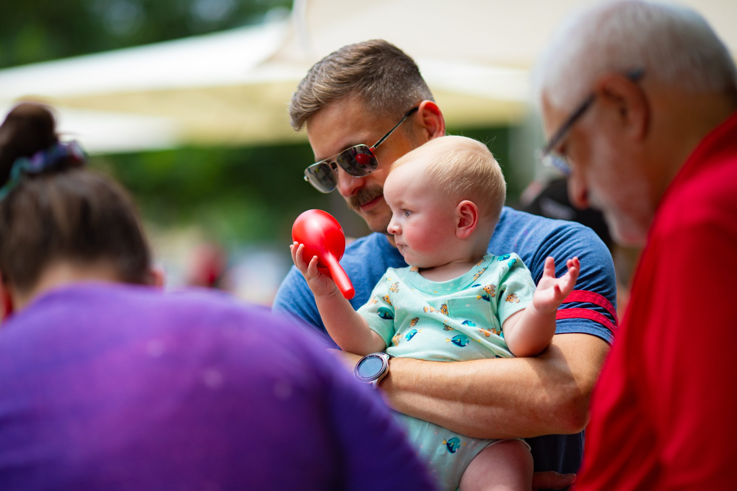 A small child held by father shaking a maraca